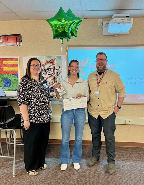 A student holds a certificate and smiles between two staff members, with green balloons in a classroom setting.