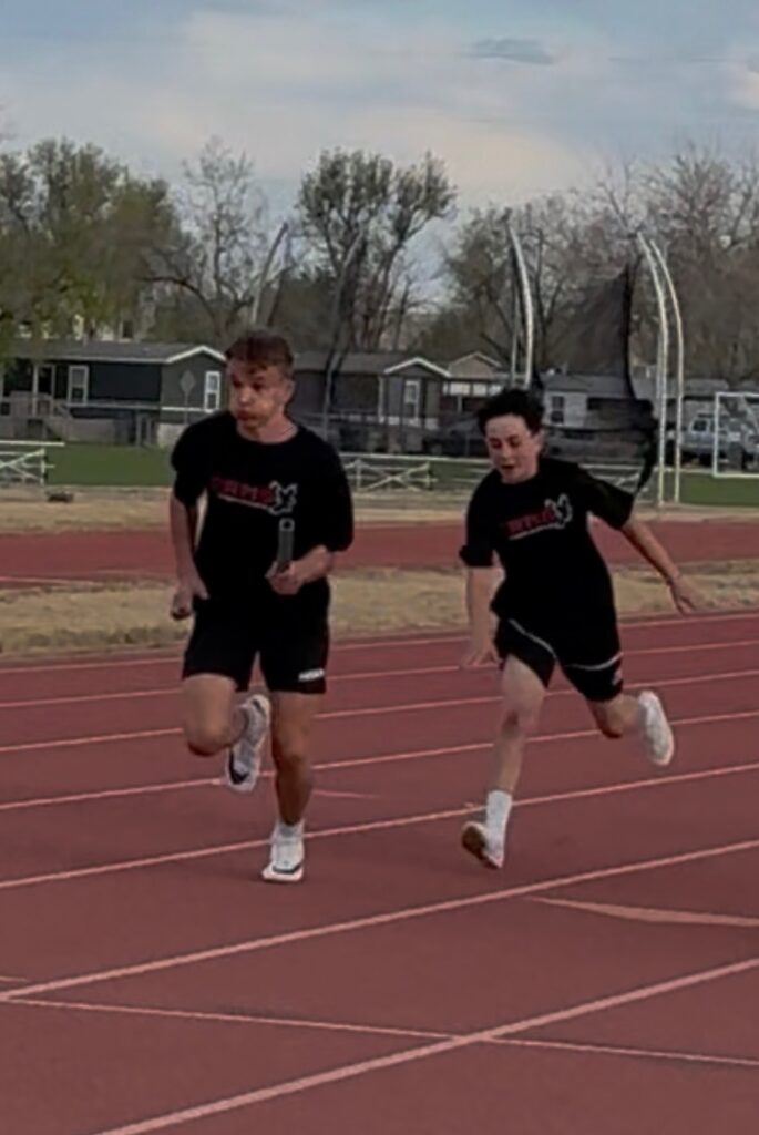 Two young athletes sprinting on a track, one holding a relay baton, with trees and buildings in the background.