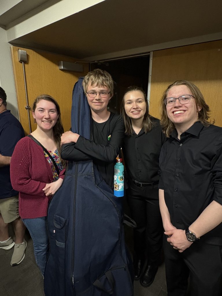 Four young people pose together, smiling, with one holding a large instrument case and a water bottle.