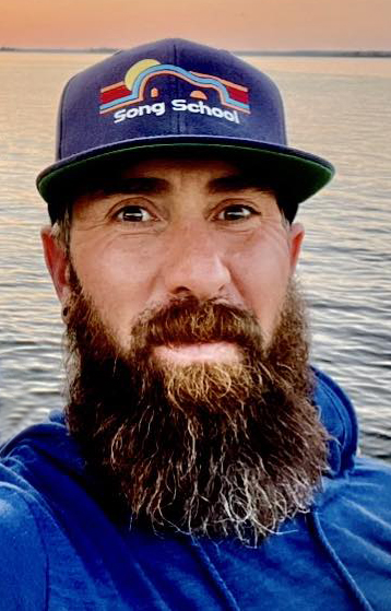 Man with a beard wearing a blue hoodie and a hat that says "Song School," with water and sunset in the background.