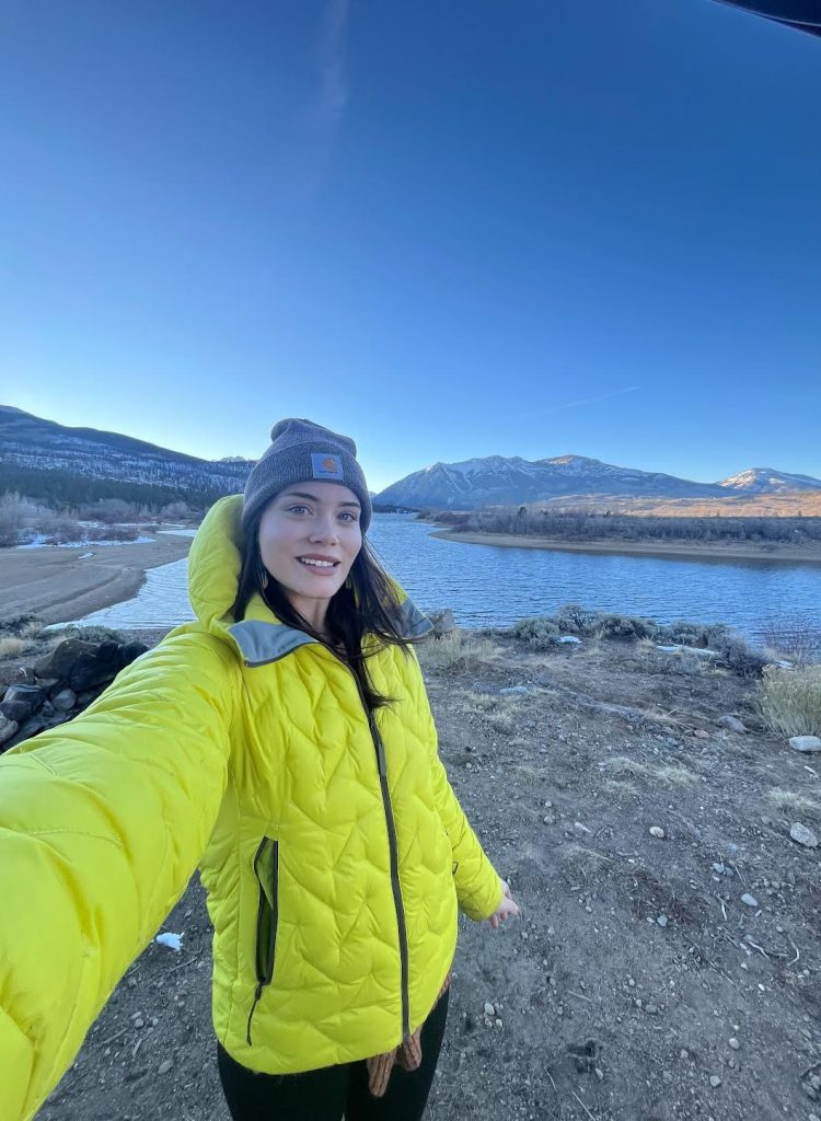 A person in a bright yellow jacket smiles for a selfie by a river with mountains in the background under a clear sky.