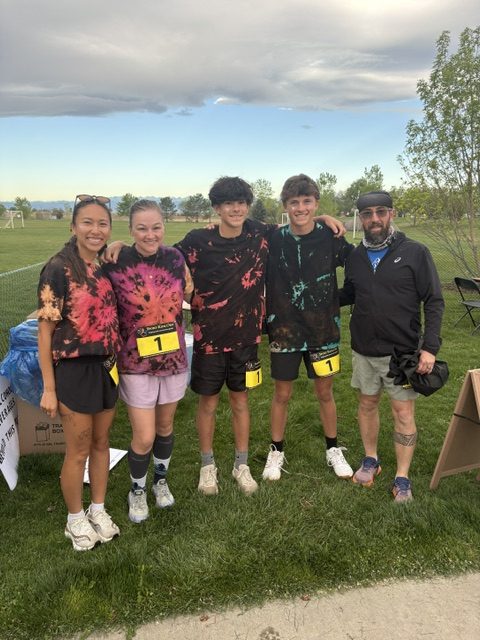 Five young people in tie-dye shirts pose together outdoors, smiling, with race numbers on their shorts.