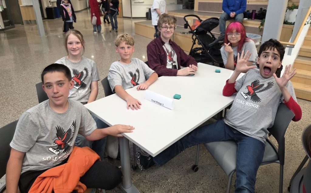 Six students in matching t-shirts sit at a table, smiling and posing, with a sign reading "Coal Ridge 2" in front.