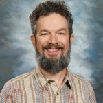Smiling man with a curly beard and plaid shirt against a cloudy backdrop.