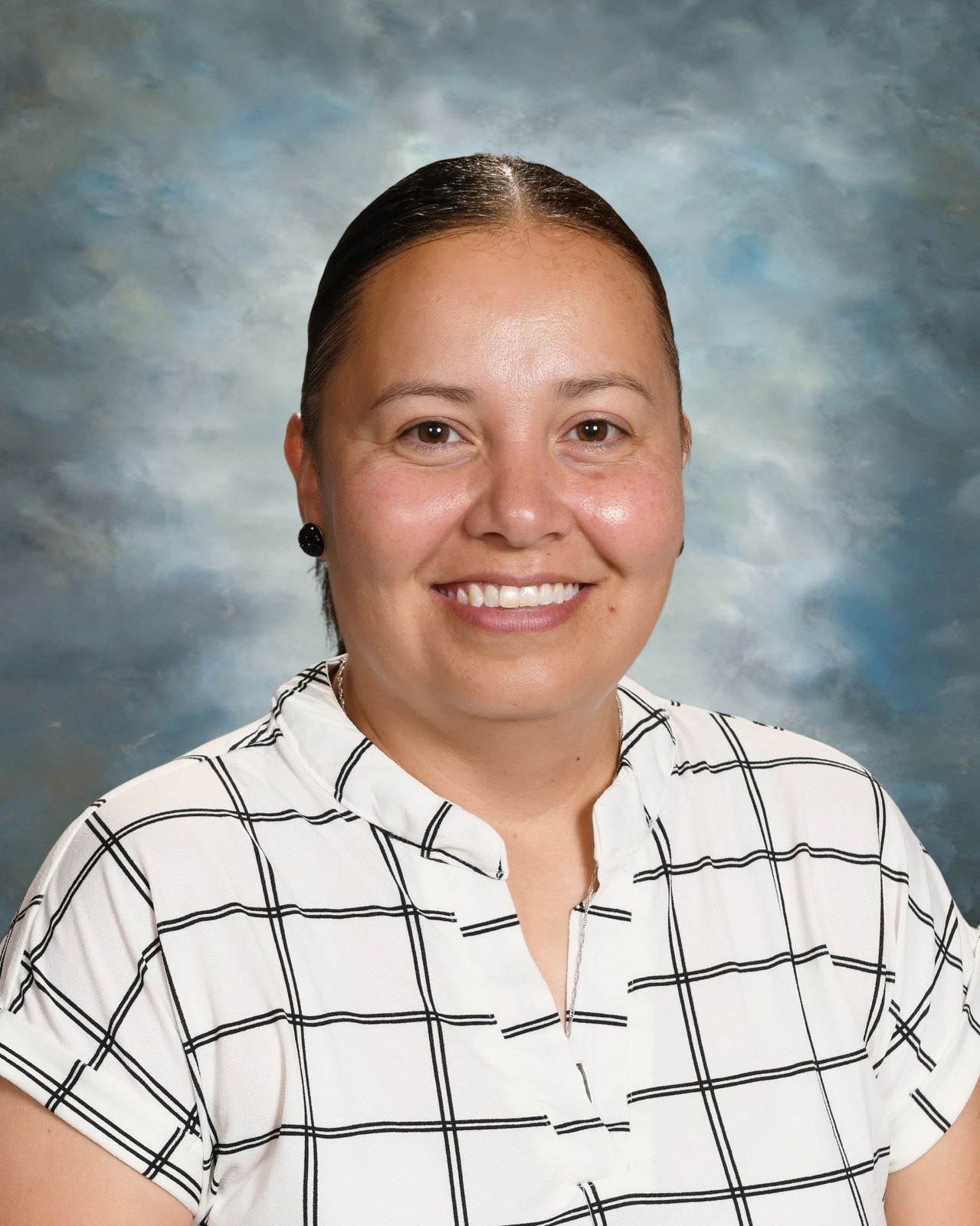 Smiling person with dark hair tied back, wearing a white plaid shirt, against a cloudy blue background.