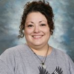 Woman smiling, wearing a gray shirt with a bird design, curly hair, and a necklace. Cloudy background.