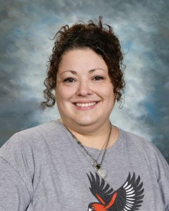 Woman smiling, wearing a gray shirt with a bird design, curly hair, and a necklace. Cloudy background.