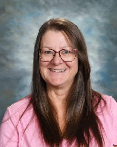 Smiling woman with long brown hair and glasses, wearing a pink shirt against a cloudy blue backdrop.