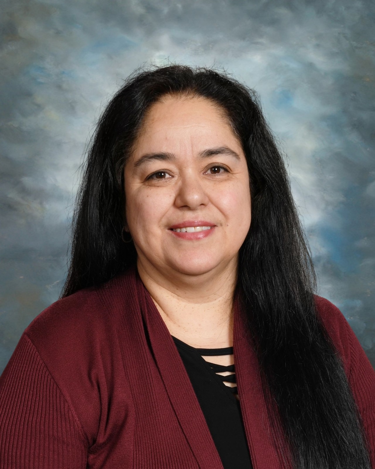 Smiling woman with long black hair wearing a maroon cardigan and black top, set against a cloudy gray background.