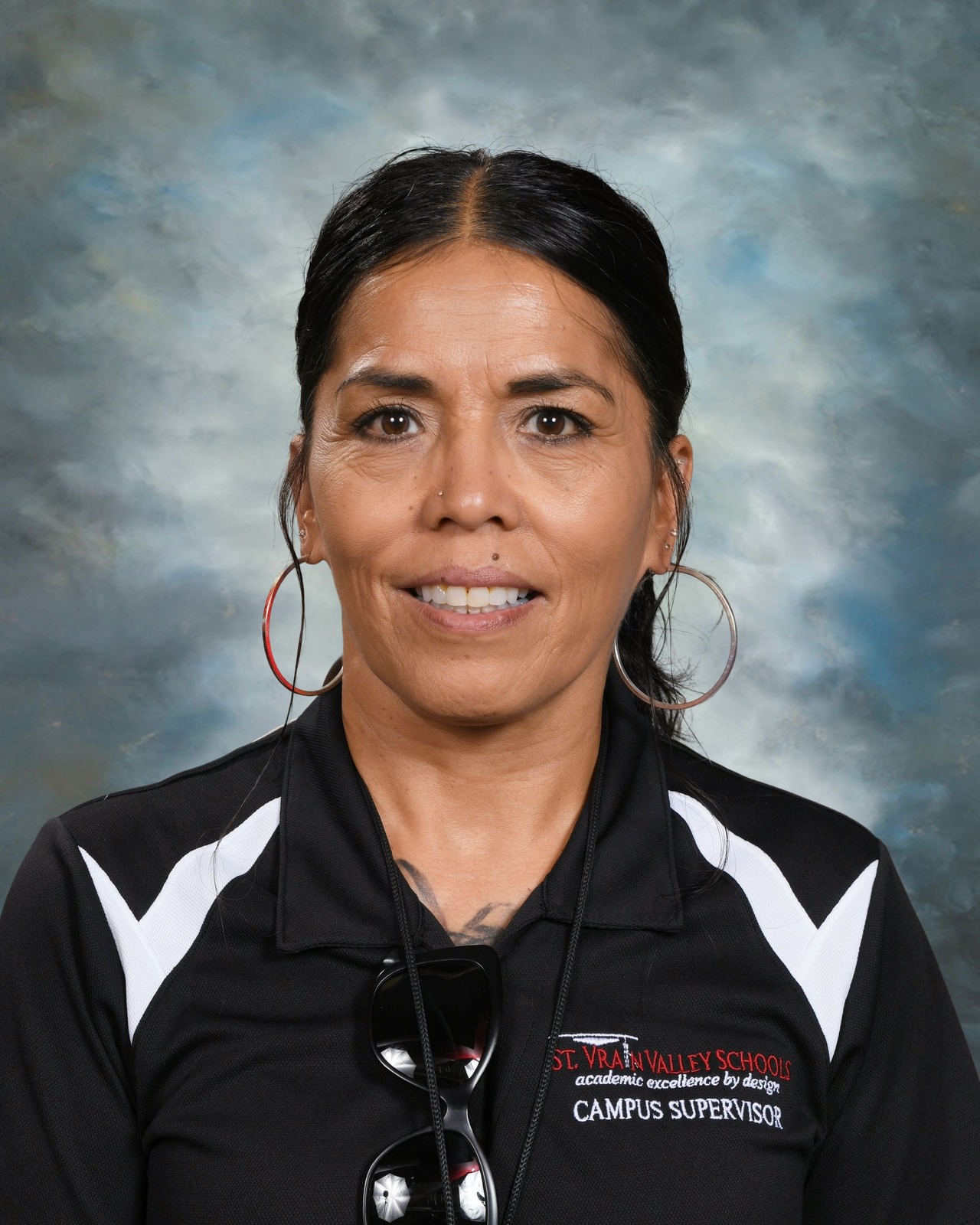 Woman with hoop earrings and black shirt with a campus supervisor logo, smiling against a cloudy background.