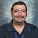 Smiling man with short hair and mustache, wearing a dark polo shirt and blue lanyard against a cloudy backdrop.