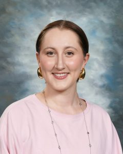 Smiling person with short hair wearing a pink shirt and gold earrings against a cloudy background.